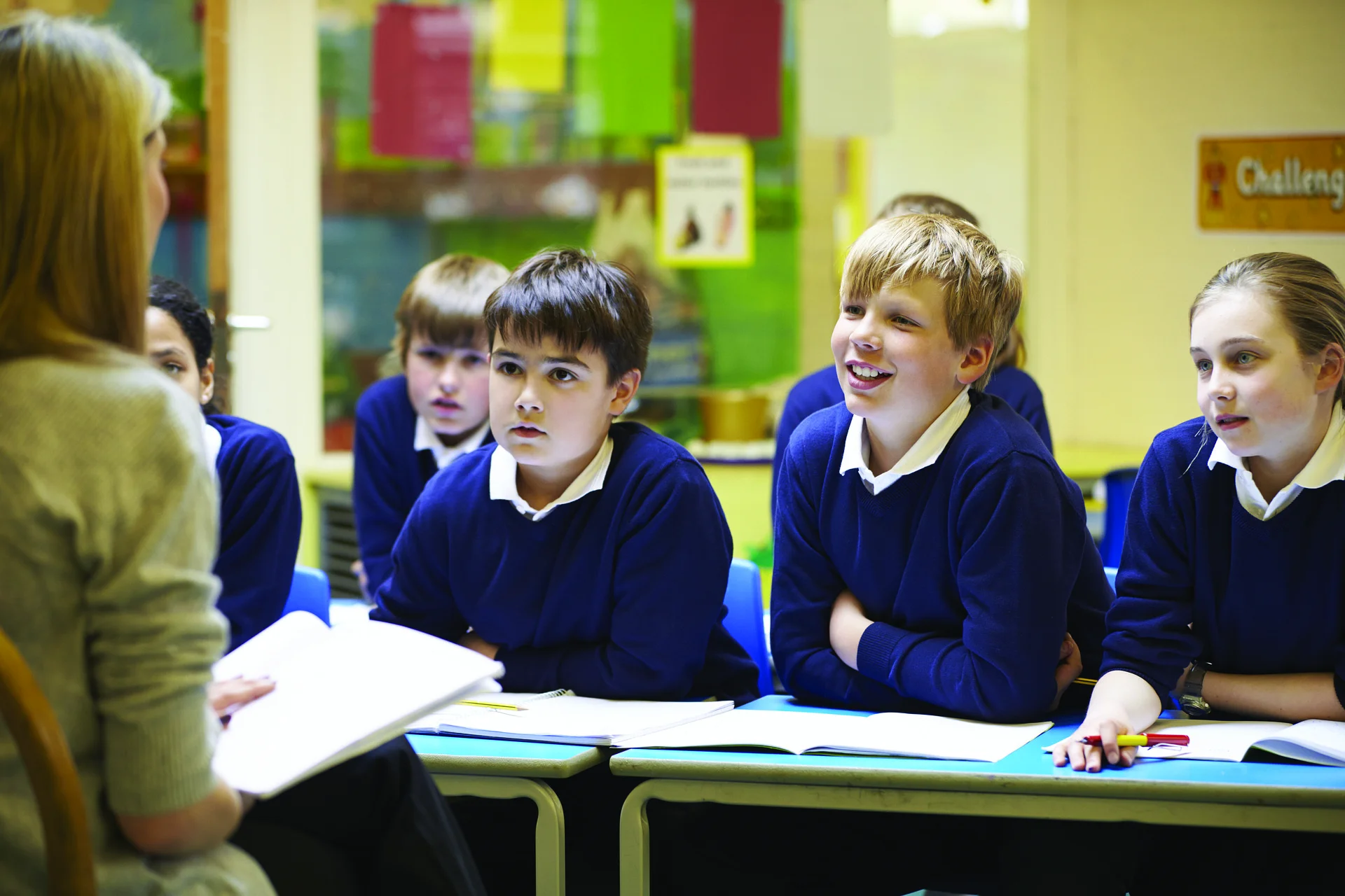 Teacher leads a small class discussion with students in blue sweaters seated at desks, notebooks open.