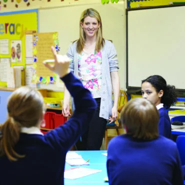 Teacher standing at the front of a colorful classroom, smiling as students raise their hands to answer a question.