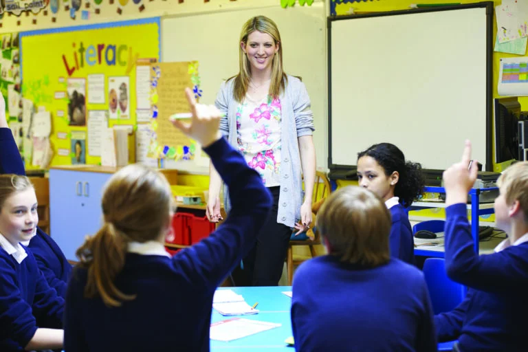 Teacher standing at the front of a colorful classroom, smiling as students raise their hands to answer a question.