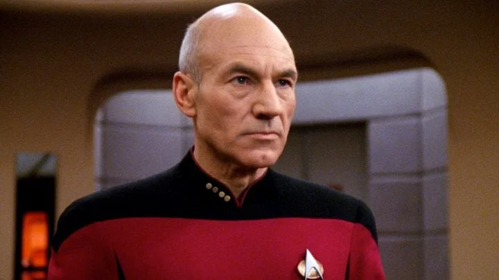 Bald man in a red and black Starfleet uniform on a starship bridge with a serious expression.