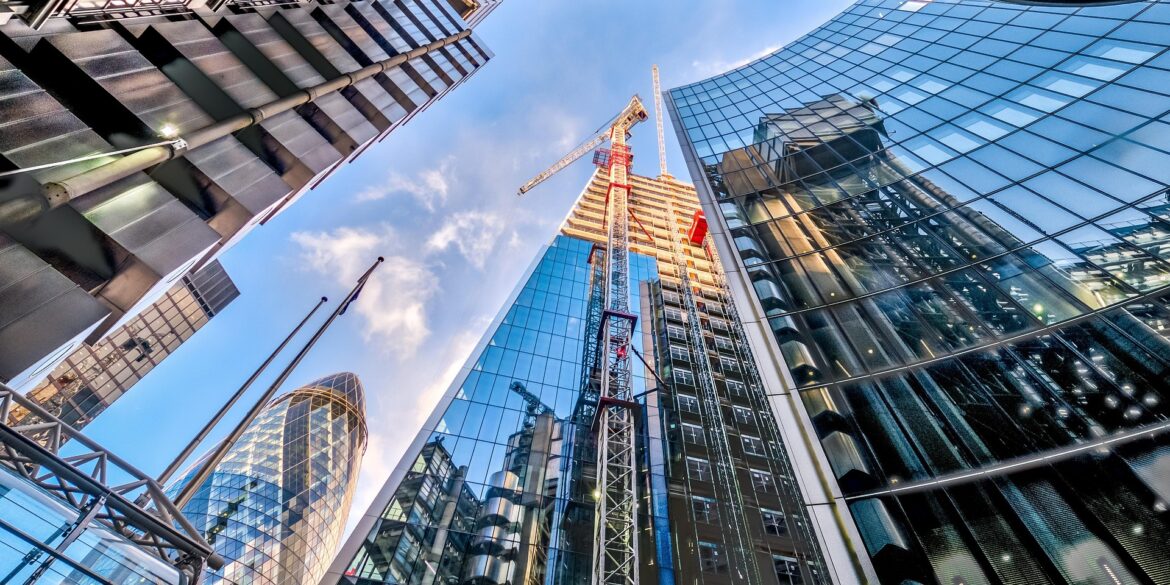 Construction crane rises between modern glass skyscrapers under a blue sky, in a metropolitan area.