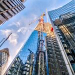 Construction crane rises between modern glass skyscrapers under a blue sky, in a metropolitan area.