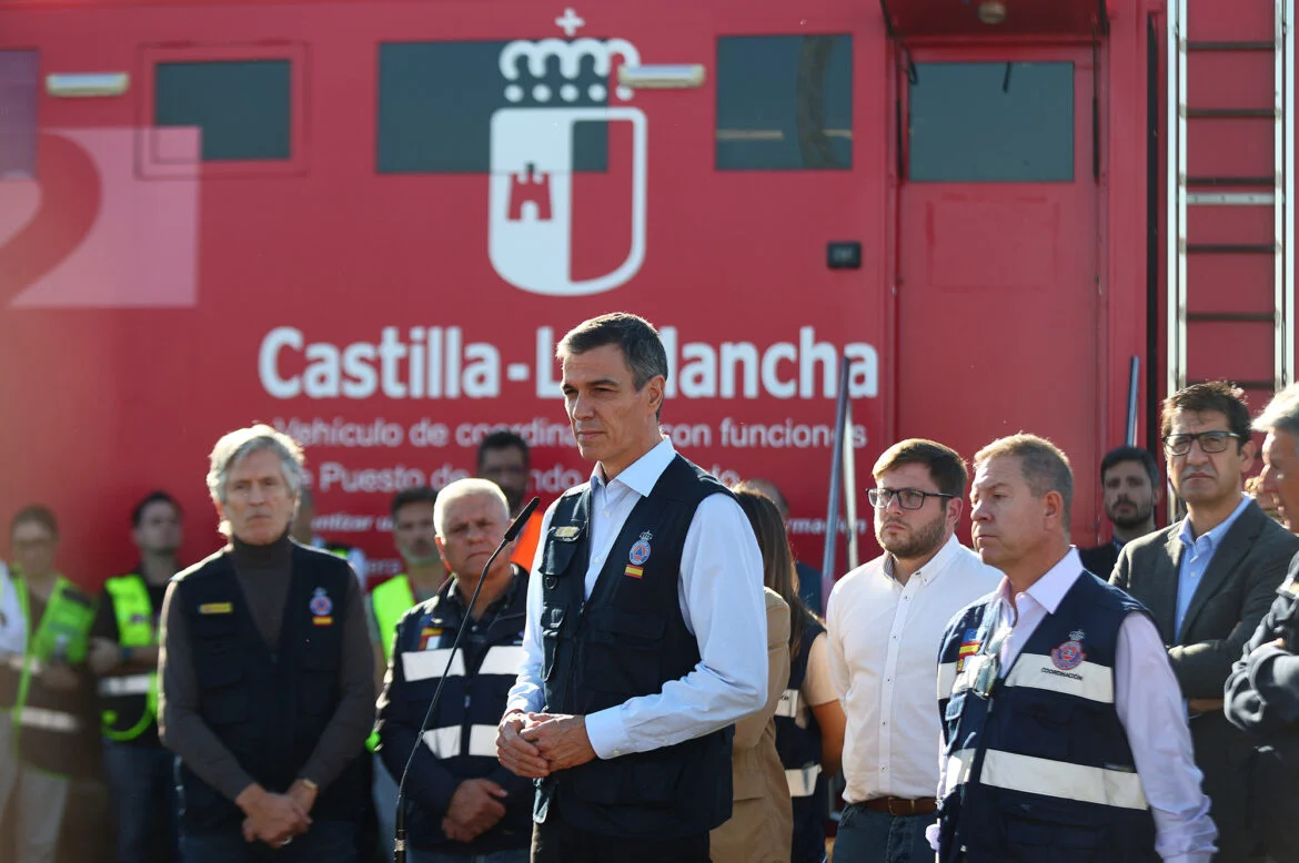 Man in a vest speaks into a mic at a briefing, in front of a red Castilla-La Mancha vehicle with officials and emergency responders nearby.