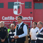 Man in a vest speaks into a mic at a briefing, in front of a red Castilla-La Mancha vehicle with officials and emergency responders nearby.