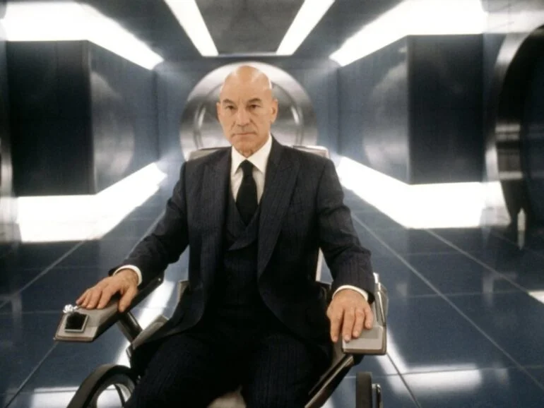 Bald man in a dark pinstripe suit sits in a futuristic wheelchair in a shiny sci-fi hallway.