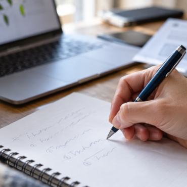 Person writes in a spiral notebook at a wooden desk, with a blue floral mug, laptop, and papers nearby.