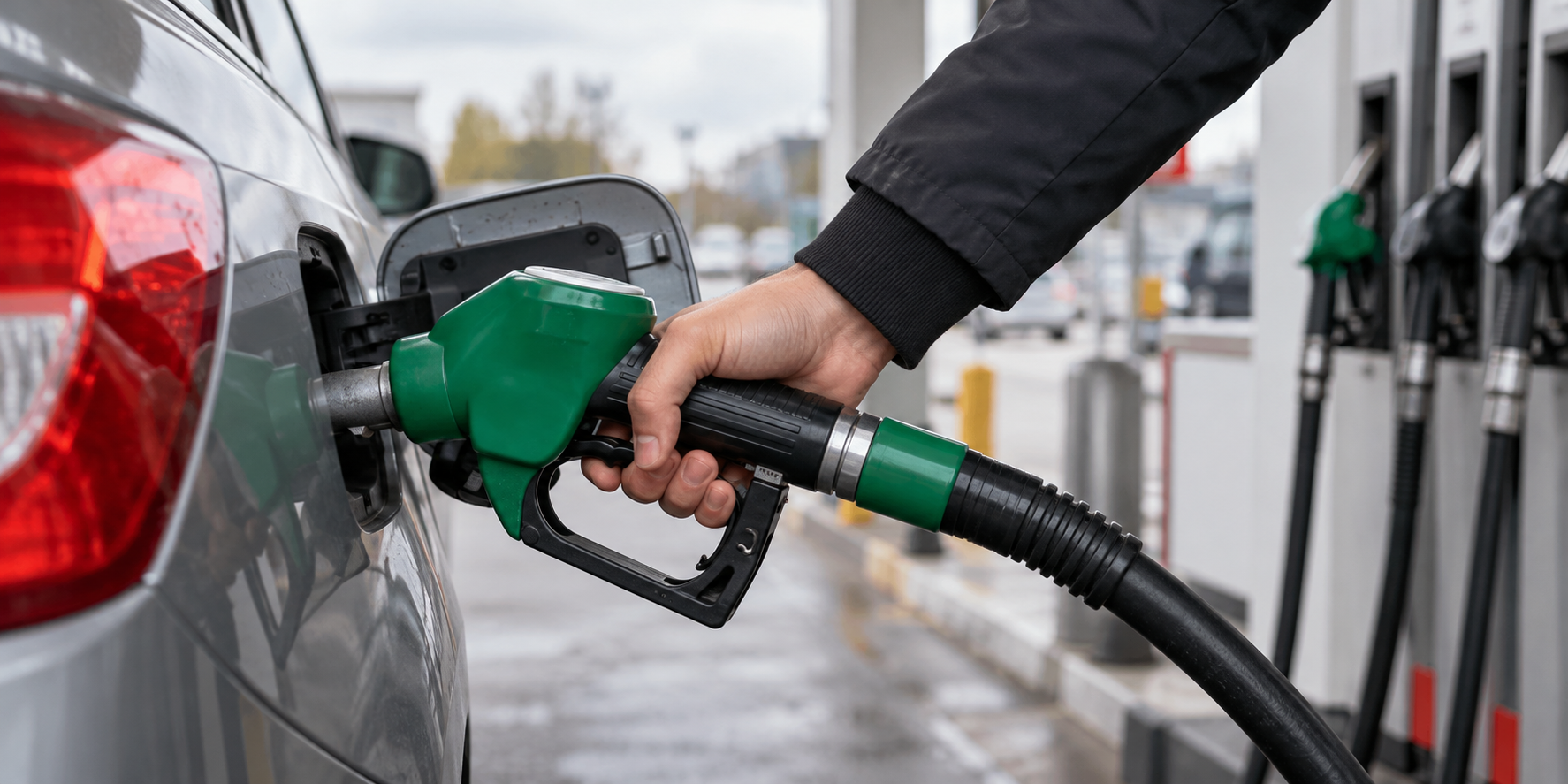 Person fueling a car at a gas station, holding a green nozzle to the fuel inlet of the car's side.