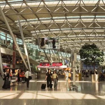 Spacious airport concourse with a glass and steel roof, travelers pulling luggage and walking near shops and signs, including a Starbucks sign on the left.
