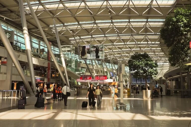 Spacious airport concourse with a glass and steel roof, travelers pulling luggage and walking near shops and signs, including a Starbucks sign on the left.