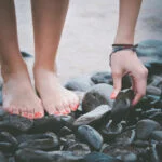 Barefeet on dark pebbles at the water's edge as a hand selects a smooth rock surface.