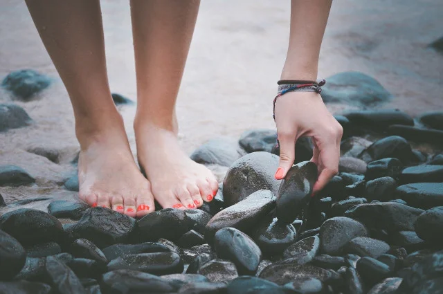 Barefeet on dark pebbles at the water's edge as a hand selects a smooth rock surface.
