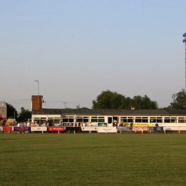 Soccer field with a player kicking the ball near the left foreground; banners and a long sideline stand in the background under a clear sky.