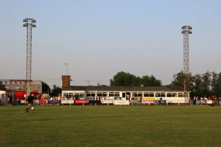 Soccer field with a player kicking the ball near the left foreground; banners and a long sideline stand in the background under a clear sky.