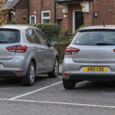 Two silver hatchback cars parked side by side in a residential street, rear ends facing the camera, with license plates AB12 CDE visible in a brick townhouse setting.
