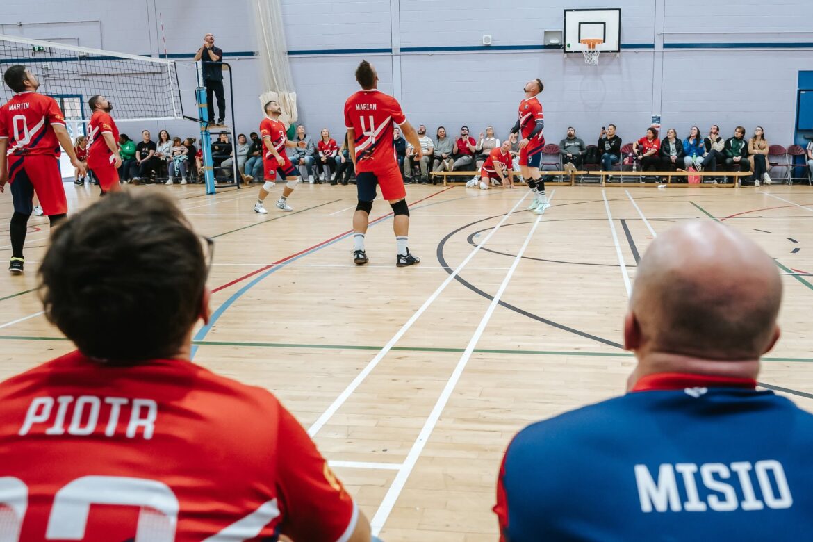 Indoor volleyball game with players in red jerseys; view from behind two teammates as they watch the action near the net, with spectators along the wall and a referee on a chair in the background, gymnasium setting.