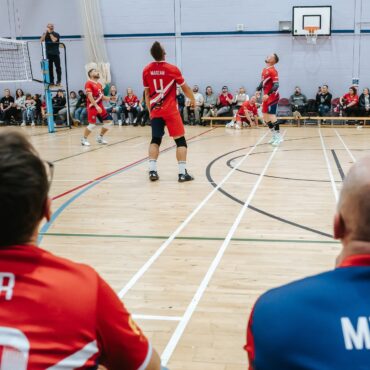 Indoor volleyball game with players in red jerseys; view from behind two teammates as they watch the action near the net, with spectators along the wall and a referee on a chair in the background, gymnasium setting.