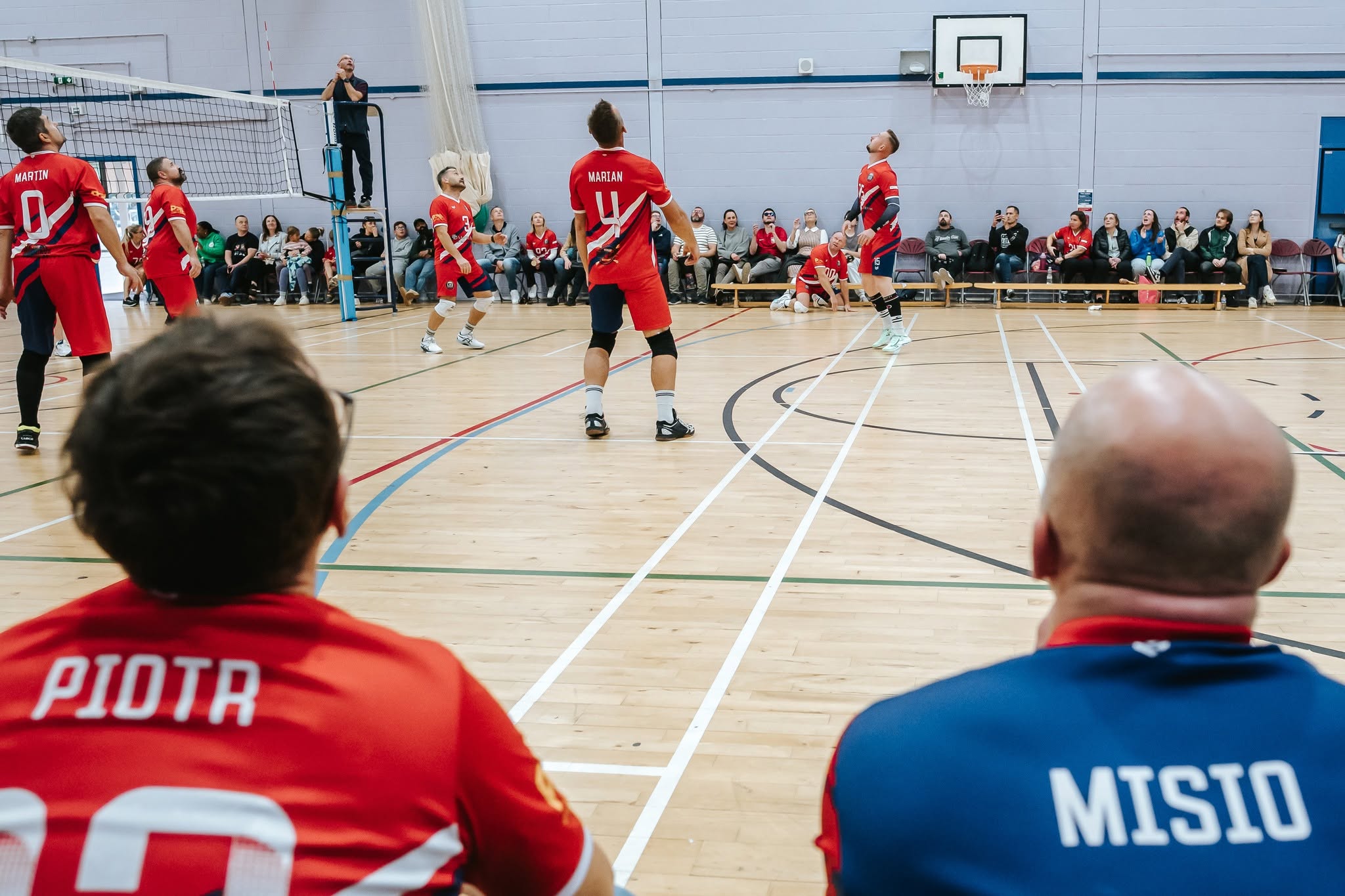 Indoor volleyball game with players in red jerseys; view from behind two teammates as they watch the action near the net, with spectators along the wall and a referee on a chair in the background, gymnasium setting.