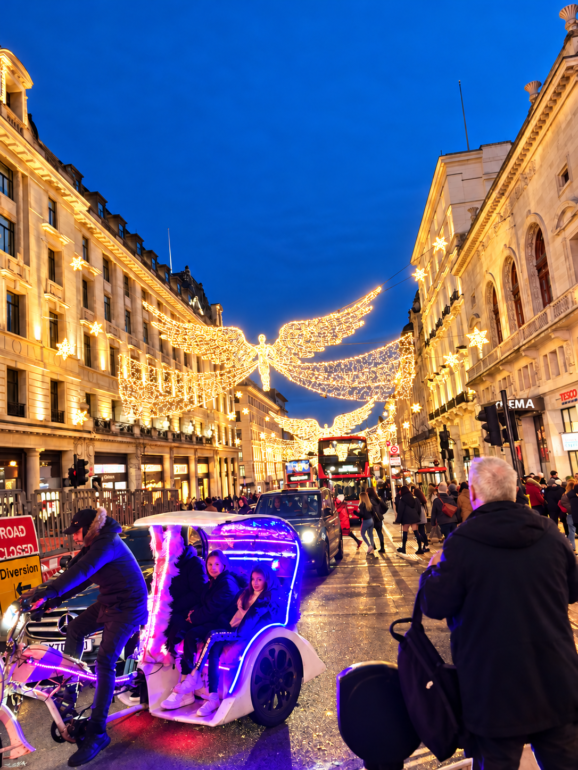 Night street lit with golden angel decorations; pedestrians bustle as a purple-lit pedicab carries three children nearby.