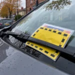 Yellow warning notice placed under the windshield wiper on a parked car in a wet urban street scene