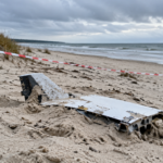 Crumbled metal debris on a sandy beach behind red-and-white caution tape, with dunes and the sea in the distance.