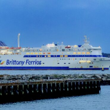 White Brittany Ferries passenger ship Barfleur on calm blue water, lined with a rocky breakwater in the foreground and a distant shoreline under a clear blue sky