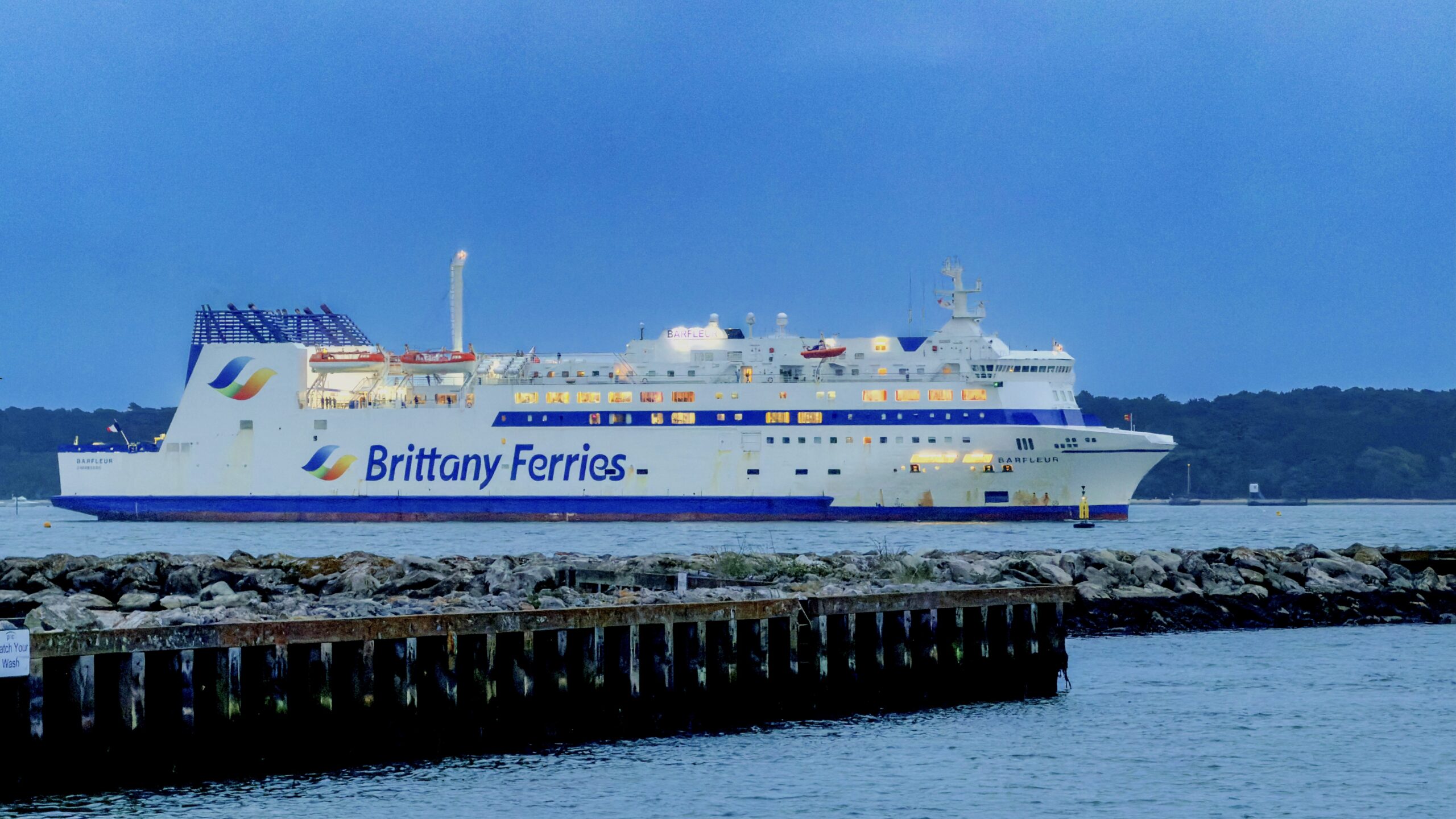White Brittany Ferries passenger ship Barfleur on calm blue water, lined with a rocky breakwater in the foreground and a distant shoreline under a clear blue sky