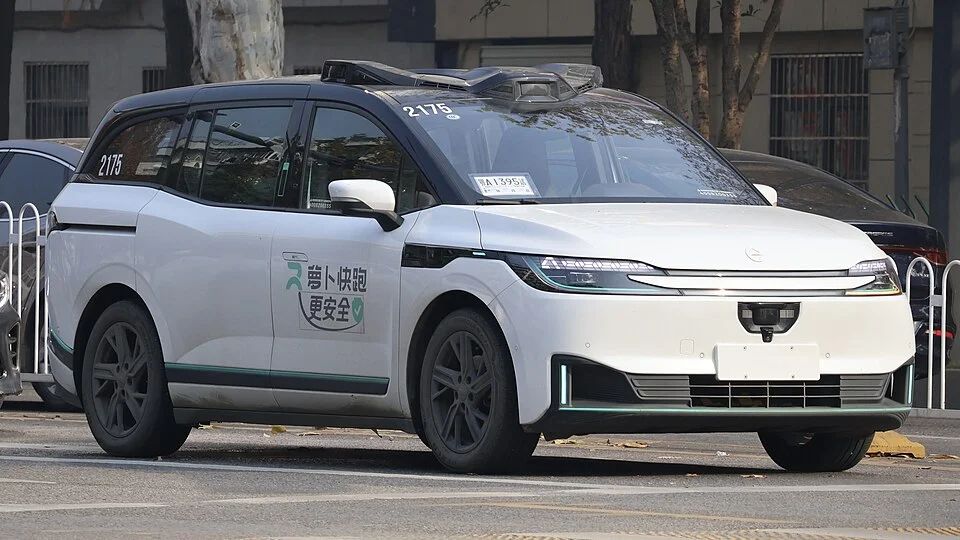 Self-driving white minivan taxi on a city street, marked with number 2175 and safety branding on the side.