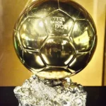 Gold Ballon d'Or trophy on a textured crystal-like base, with a plaque reading 'Lionel Messi' in front.