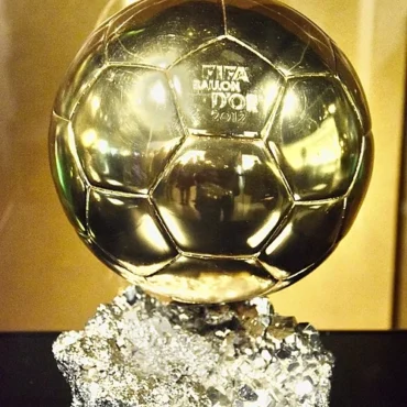 Gold Ballon d'Or trophy on a textured crystal-like base, with a plaque reading 'Lionel Messi' in front.