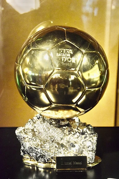 Gold Ballon d'Or trophy on a textured crystal-like base, with a plaque reading 'Lionel Messi' in front.