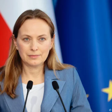 Woman in a blue blazer speaks at a press conference, with red and EU flags in the background.