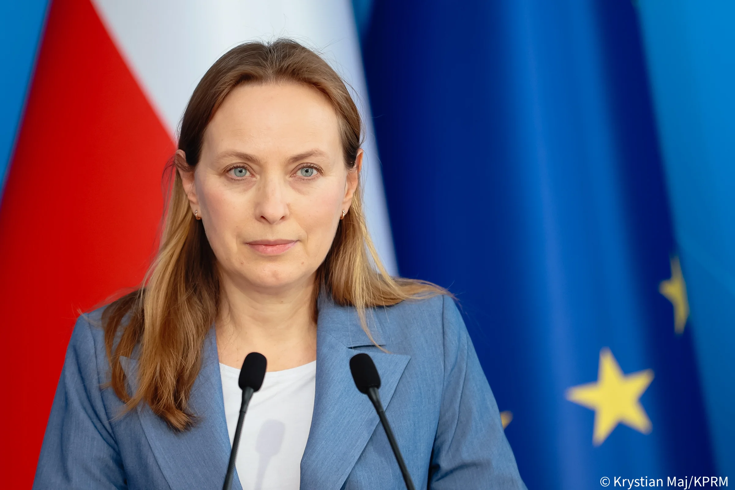 Woman in a blue blazer speaks at a press conference, with red and EU flags in the background.