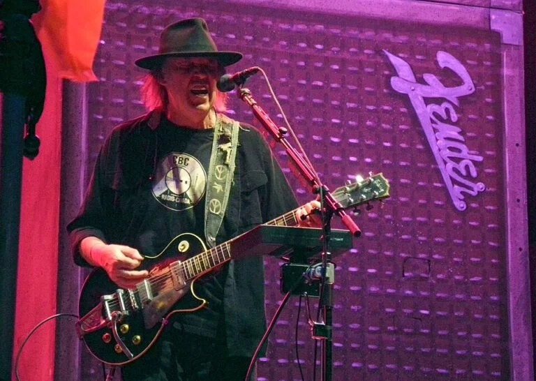 Male guitarist wearing a hat, singing into a microphone while playing a black Les Paul–style guitar on stage with purple lighting.