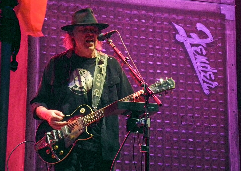 Male guitarist wearing a hat, singing into a microphone while playing a black Les Paul–style guitar on stage with purple lighting.