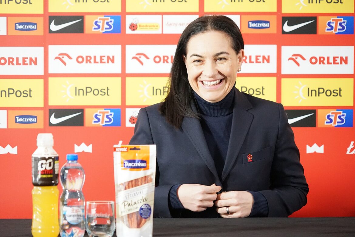 Smiling woman in a dark blazer at a press conference table, with sponsor logos on a colorful backdrop; bottles and a snack package visible on the table.