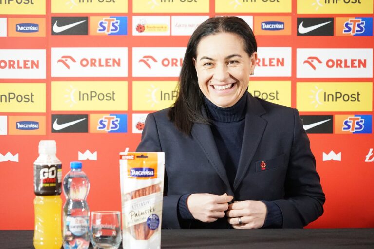 Smiling woman in a dark blazer at a press conference table, with sponsor logos on a colorful backdrop; bottles and a snack package visible on the table.
