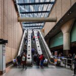 Busy indoor transit station with people ascending and descending escalators beside brick walls and a glass ceiling.