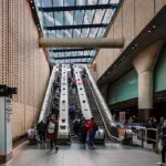 Busy indoor transit station with people ascending and descending escalators beside brick walls and a glass ceiling.