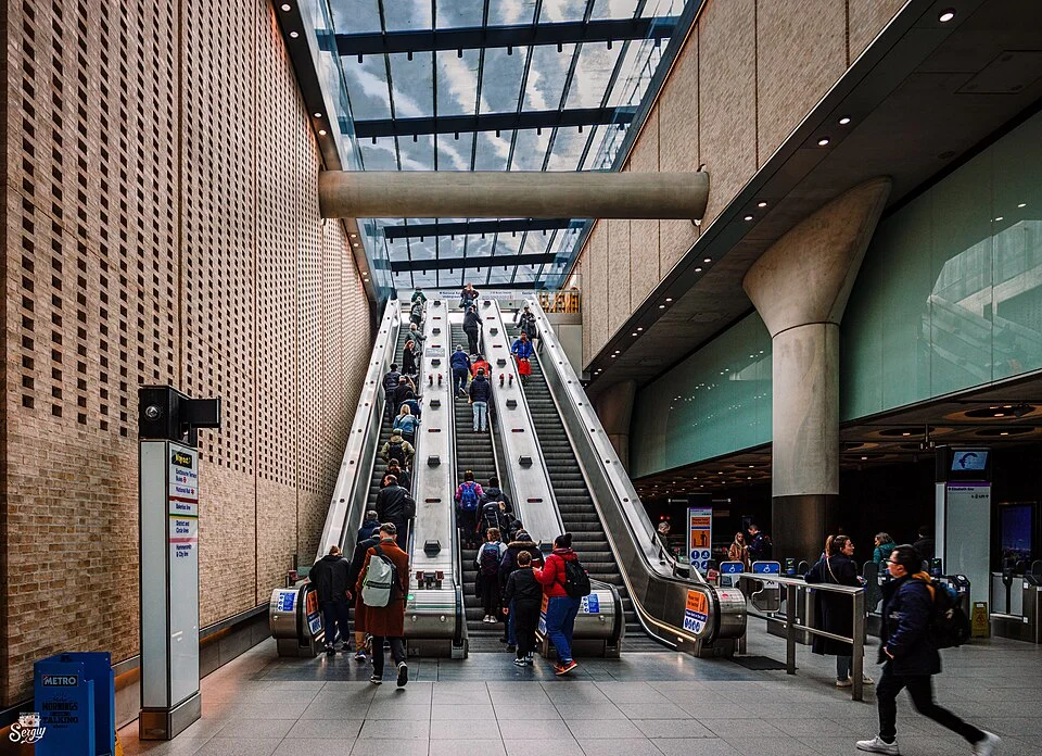 Busy indoor transit station with people ascending and descending escalators beside brick walls and a glass ceiling.