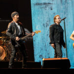 Rock band performing on stage: guitarist in leather jacket center, singer with mic, bassist on the right, drummer behind them on the left with a full drum kit against a blue textured backdrop.