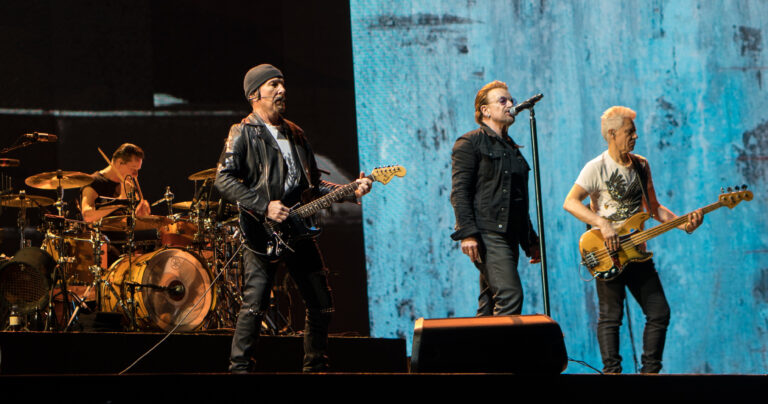 Rock band performing on stage: guitarist in leather jacket center, singer with mic, bassist on the right, drummer behind them on the left with a full drum kit against a blue textured backdrop.