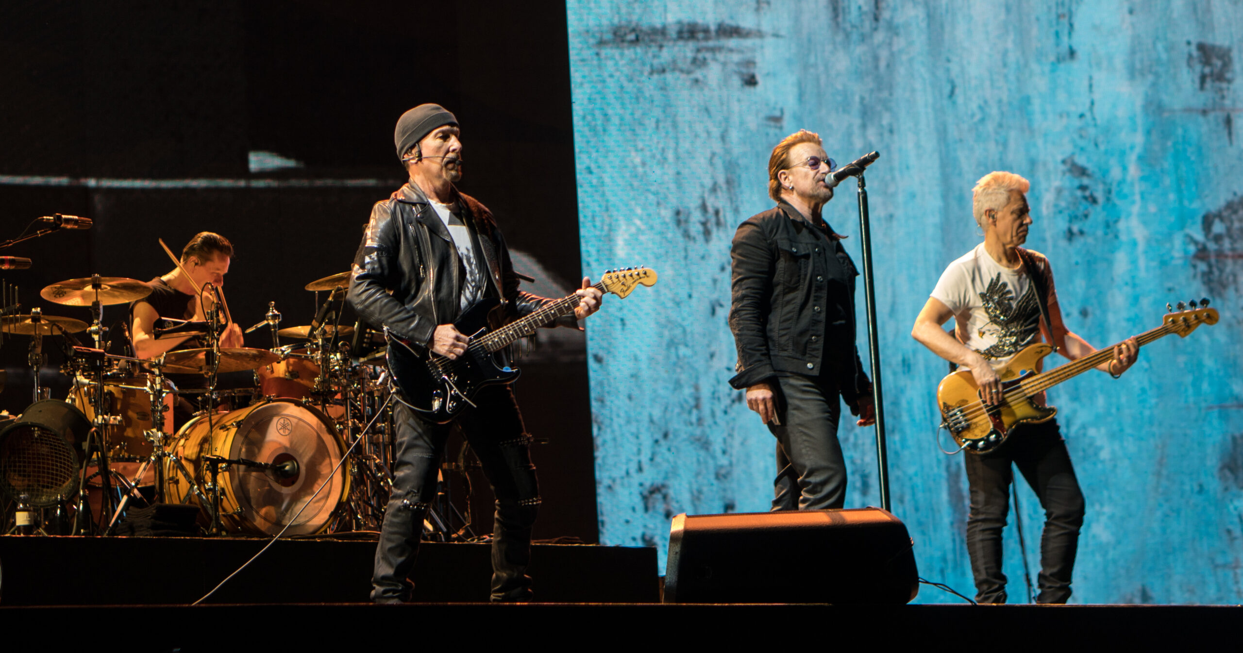 Rock band performing on stage: guitarist in leather jacket center, singer with mic, bassist on the right, drummer behind them on the left with a full drum kit against a blue textured backdrop.