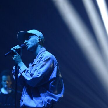 Male singer wearing a cap performs into a microphone on a blue-lit stage, with a keyboardist in the background.