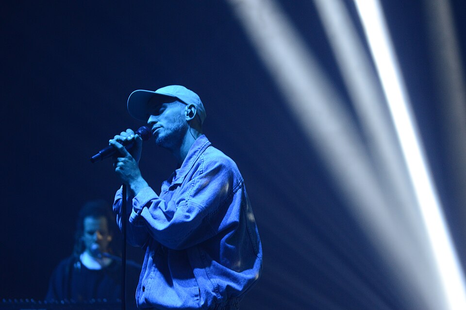 Male singer wearing a cap performs into a microphone on a blue-lit stage, with a keyboardist in the background.