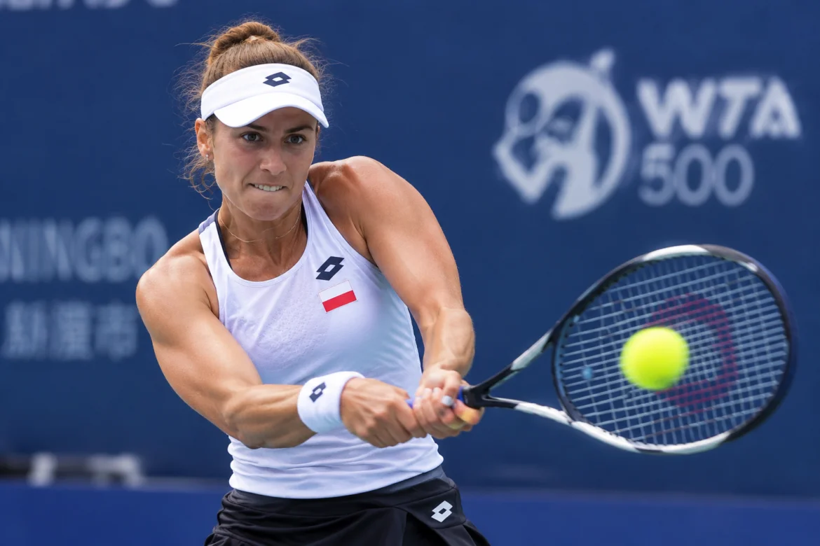 Female tennis player in a white tank top and visor hits a two-handed backhand volley toward the ball during a match.