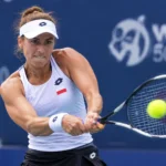 Female tennis player in a white tank top and visor hits a two-handed backhand volley toward the ball during a match.