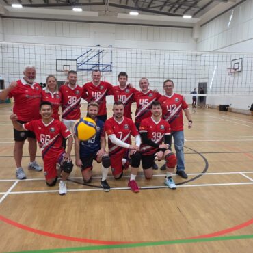 Group of volleyball players in red uniforms posing for a team photo on an indoor court with a net behind them.