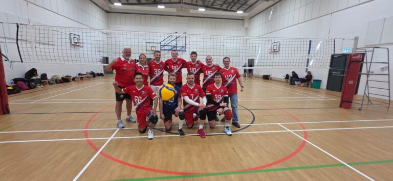 Group of volleyball players in red uniforms posing for a team photo on an indoor court with a net behind them.