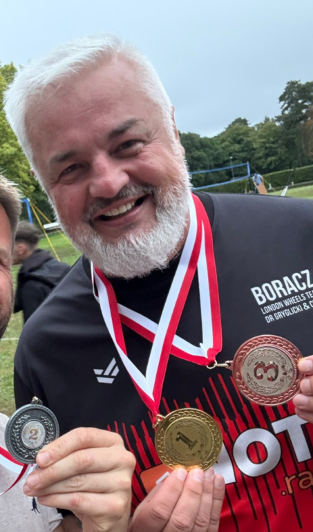 Tomasz "Boracz" Borowski - NOTE.radio - Polskie radio w UK | Polskie radio w Londynie Smiling older man with a white beard wearing a black sports jersey, displaying gold, silver, and bronze medals on red and white ribbons outdoors in a park.