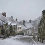 Snowy historic street: stone houses with snow-covered roofs on left, a ancient stone wall on right, light snowfall over cobbled road.